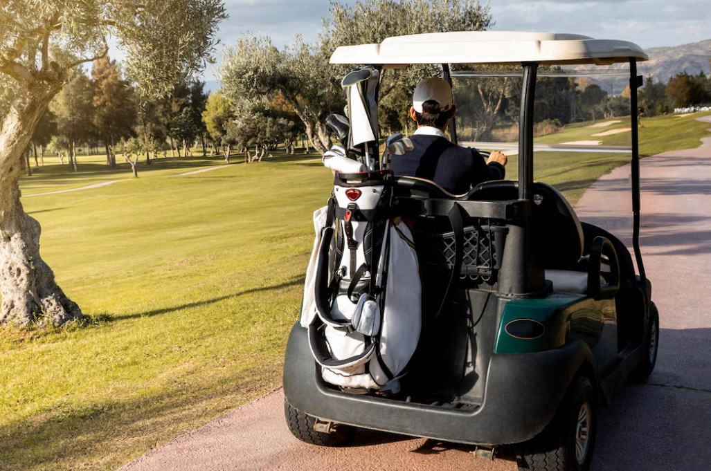 man in white hat and blue sweater riding on a golf cart on the road among the fields with trees